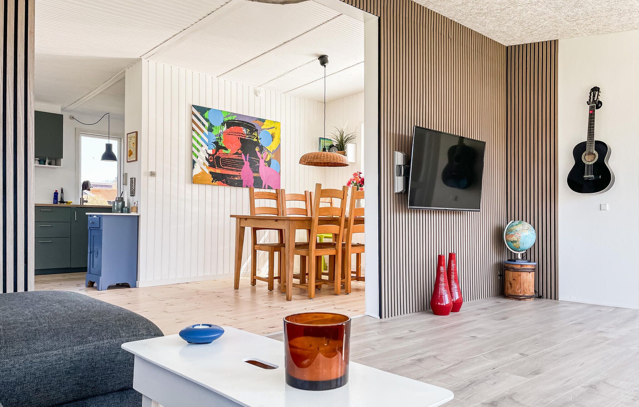 A modern living room with a dining area, a television mounted on the wall, and a guitar.