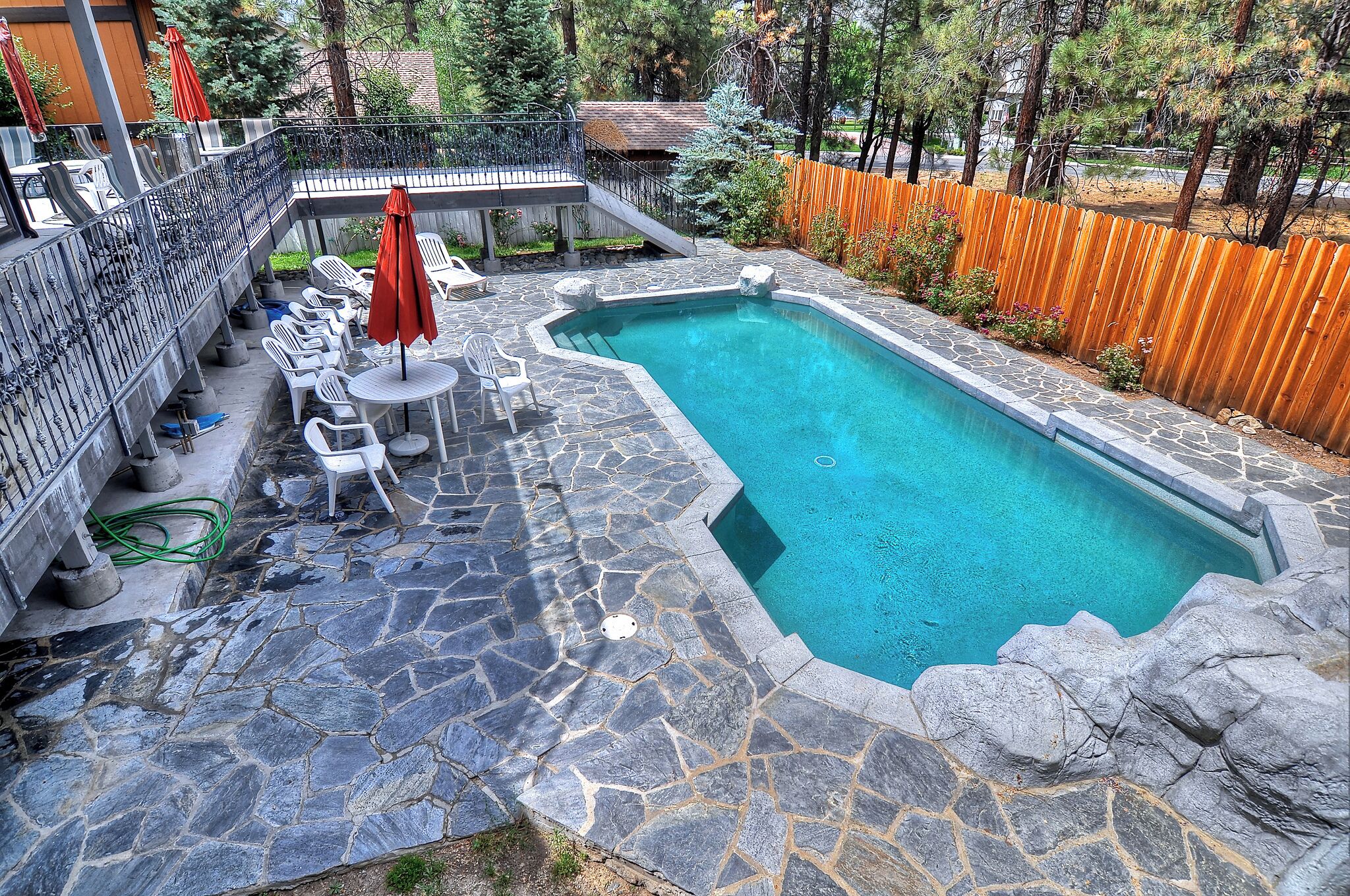 A swimming pool with stone flooring, surrounded by white chairs and red umbrellas, and a wooden fence.