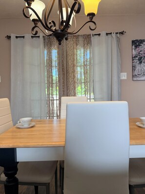 Un coin repas avec une table en bois, des chaises blanches, un lustre et une fenêtre avec des rideaux.