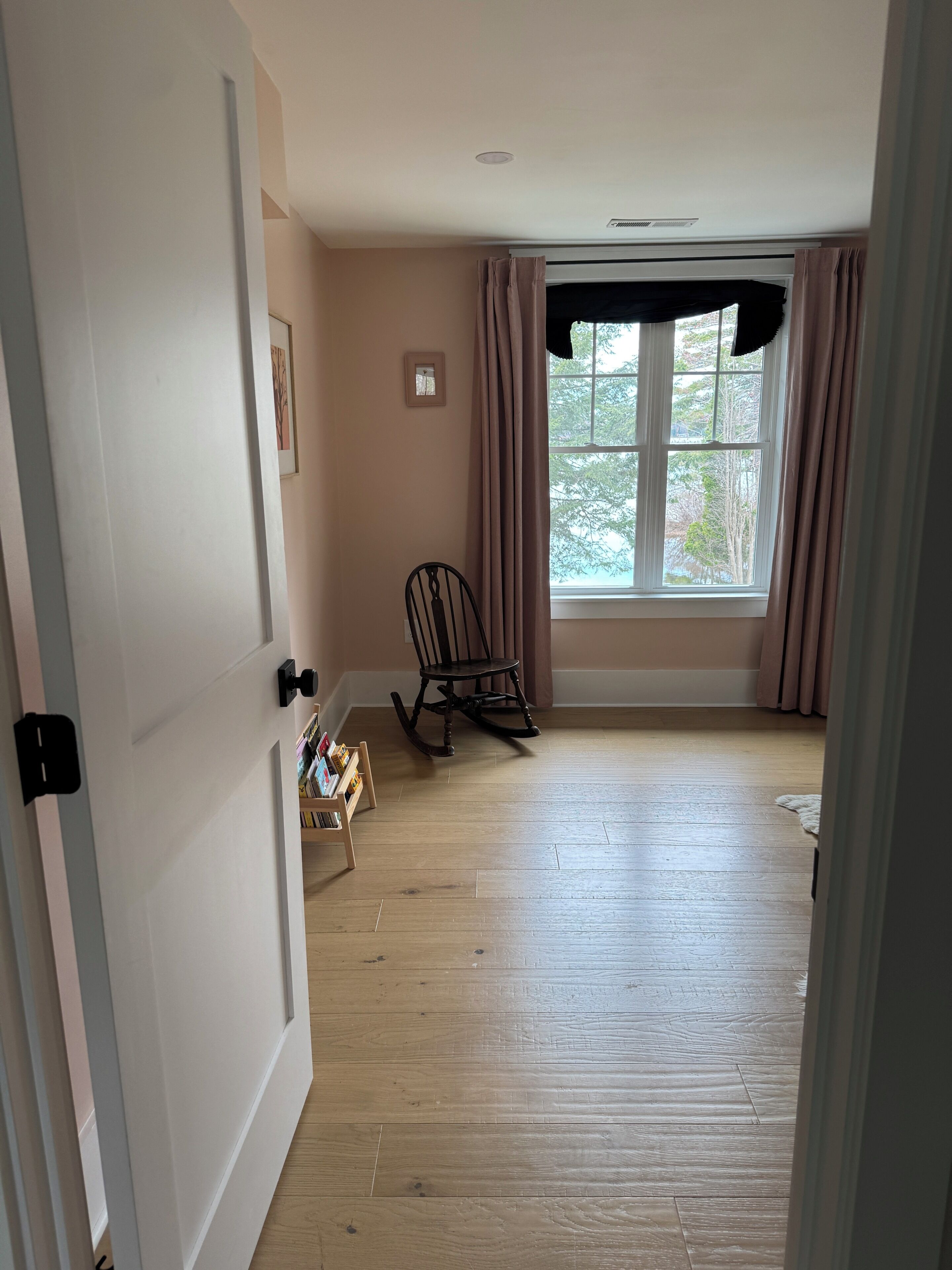 Um quarto com piso de madeira, uma cadeira de balanço, uma janela com cortinas e uma estante de livros.