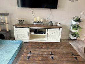 Um quarto com uma mesa de centro de madeira, um móvel branco com portas deslizantes, um arranhador para gatos, uma TV fixada na parede e um suporte de plantas com vasos identificados.