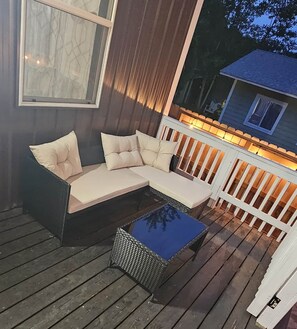 Eine Holzterrasse mit einer dunkelgrauen Couch und einem kleinen Tisch, einem weißen Geländer und einem Fenster mit Blick auf ein Haus.