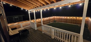 A covered outdoor deck with a wooden railing, a seating area with a table and chairs, and string lights.