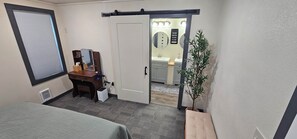 Ein Hotelzimmer mit einem Bett, einem Schreibtisch mit Computer, einer kleinen Mülltonne, einer Topfpflanze und einem durchgehenden Tür angedeuteten Badezimmer.