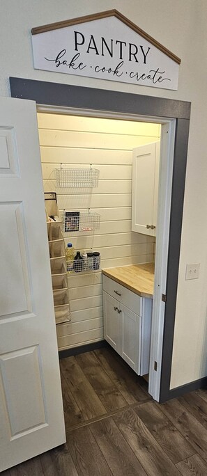 Eine Speisekammer mit einem Schild, das 'PANTRY backen kochen kreieren' aufführt, Holzschränken und Drahtkörben auf Regalen.