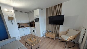 A modern living room with a sofa, a coffee table, a TV, a wicker chair, and a kitchen area with appliances. - T2 Bis Swimming pool and sea view (Agde)