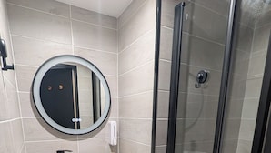 A modern bathroom with a round mirror, a white sink, a glass shower enclosure, and wooden storage shelves. - T2 Bis Swimming pool and sea view (Agde)