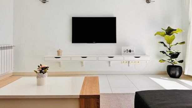 Uma sala de estar moderna com uma TV de tela plana na parede, um móvel branco embaixo, uma mesa de centro de madeira, um sofá preto e um vaso com planta.