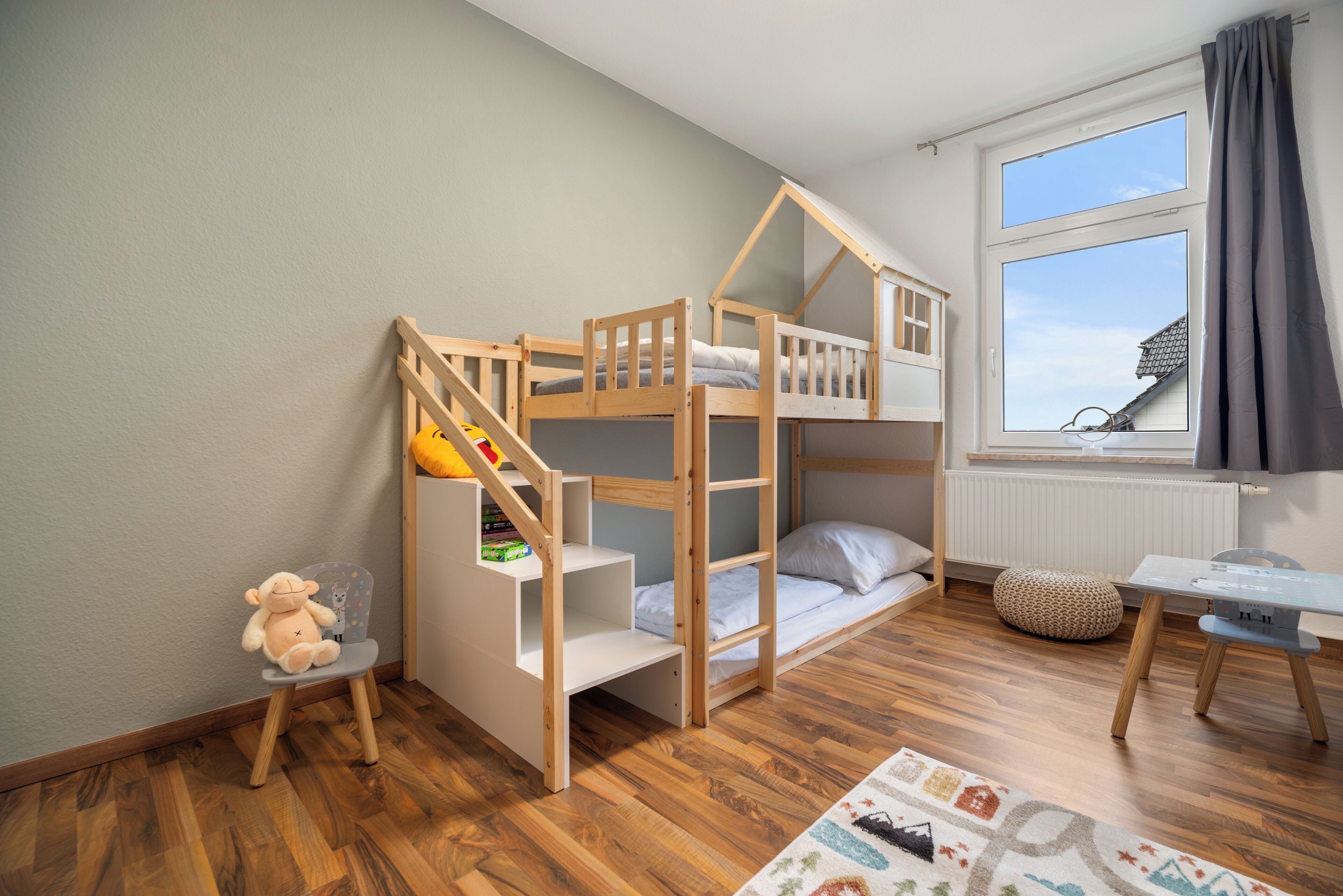 Een slaapkamer met een stapelbed, een klein bureau en stoel, een raam met gordijnen, en een pluchen speelgoed op een stoel.