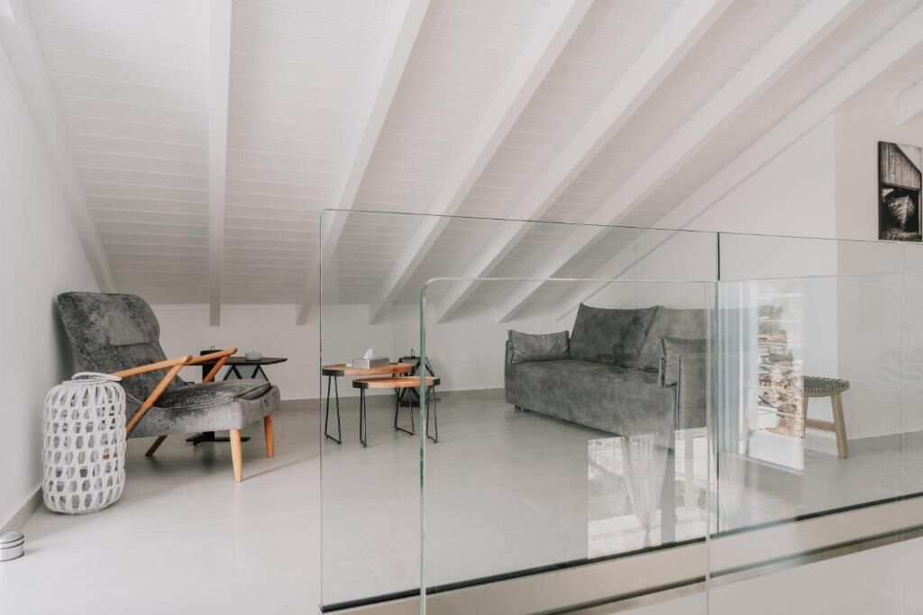 A modern loft bedroom with a glass partition, a grey sofa, a wooden chair, and a small table.