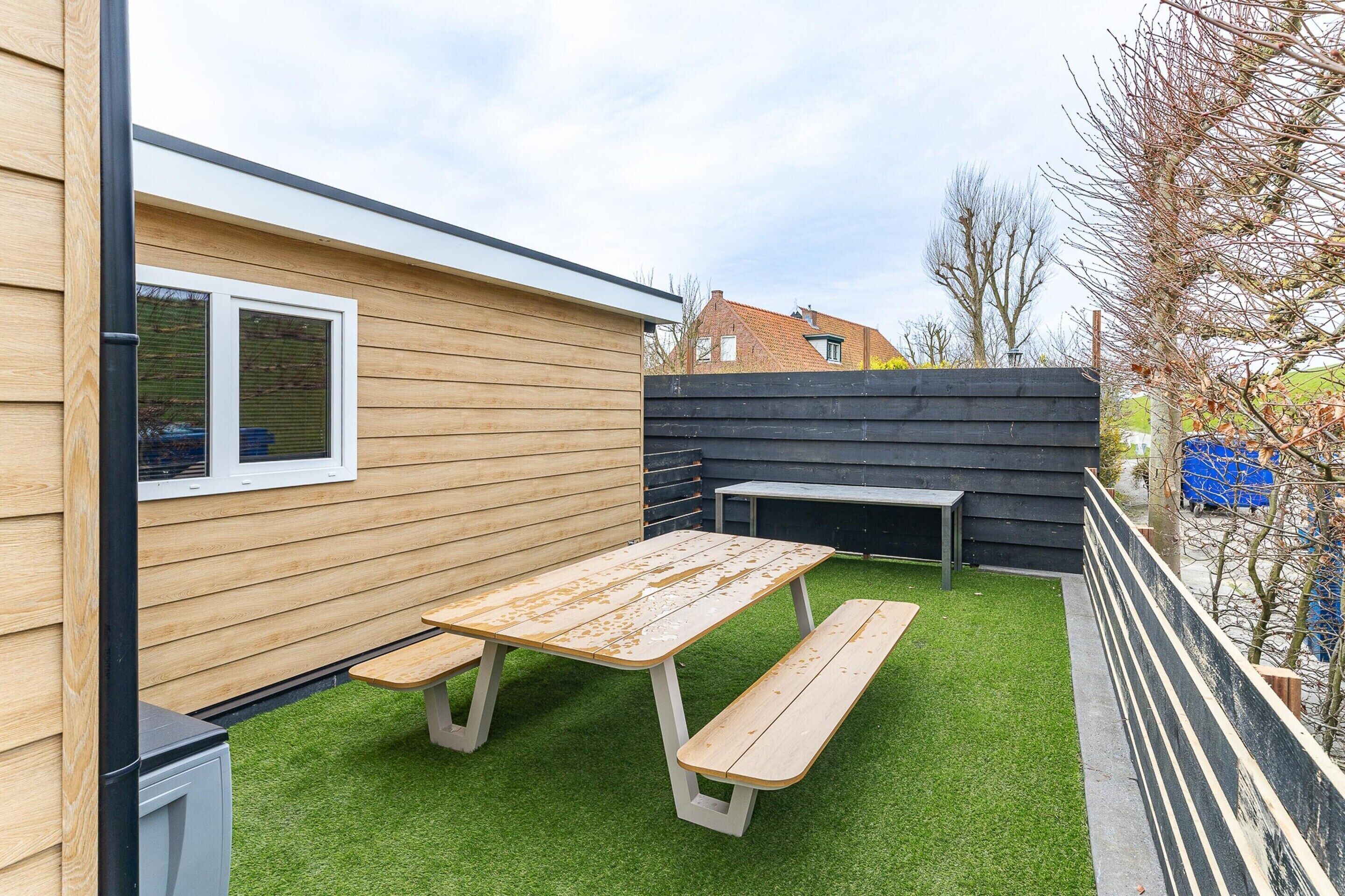 Un patio con una mesa y bancos de madera, rodeado por una valla de madera y un muro negro.