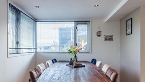 A dining area with a large wooden table, six chairs, a vase with flowers, and a window with blinds. - Serene Waterfront Escape With Sauna (Stavenisse)