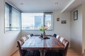 A dining area with a large wooden table, six chairs, a vase with flowers, and a window with blinds. - Serene Waterfront Escape With Sauna (Stavenisse)