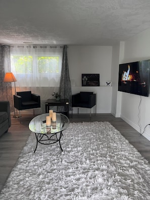 Una sala de estar moderna con una mesa de centro de vidrio, un televisor de pantalla plana, un alfombra gris y mullido, y una ventana con cortinas traslúcidas.