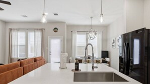 Una cocina moderna con una isla central, lavavajillas, refrigerador, luces colgantes y un área de estar con sofá.