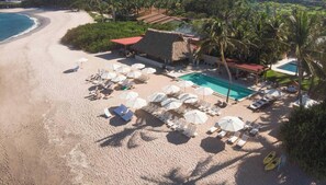 Ein Strandresort mit Pool, Liegestühlen, Sonnenschirmen und einem Gebäude mit Reetdach.