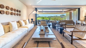 Uma sala de estar moderna com sofá, mesa de centro e cadeiras, um ventilador de teto e vista para o verde lá fora.