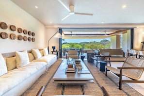 Ein modernes Wohnzimmer mit Sofa, Couchtisch und Sesseln, einem Deckenventilator und Blick auf Grünflächen.