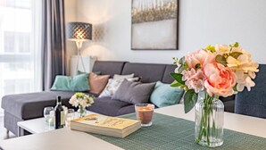 Ein modernes Wohnzimmer mit einer Couch, einem Glastisch mit einem Buch und einem Getränk sowie einer Vase mit Blumen.