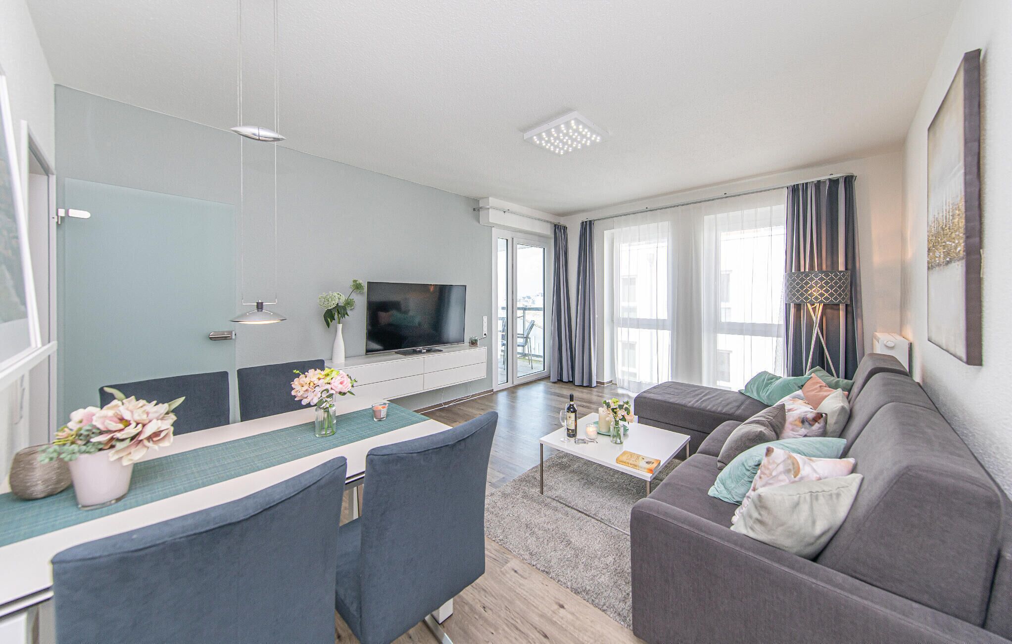 A modern living room with a flat-screen TV, grey sofa, dining table, and chairs.