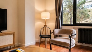 Una habitación con televisión sobre un soporte de madera, una lámpara, una silla y vistas a árboles a través de una ventana.