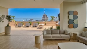 A modern living room with a sofa, coffee table, and chairs, a balcony with outdoor furniture, and a city skyline view. - Niza City Tower Downtownsd Pool Gym Balcony (Santo Domingo)