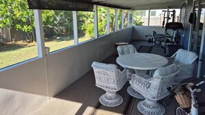 En overdækket terrasse med et rundt bord og hvide fletstole, en cykel og udsigt til en have.
