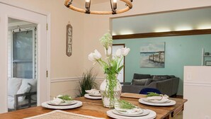 A dining area with a wooden table set for four, a vase with flowers, and a painting on the wall. - Beach Oasis, 4BR, Sleeps 12, Mins to Arrowhead (Raytown)