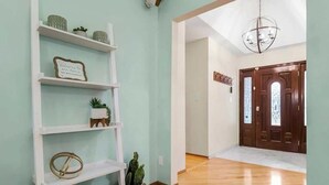 A hallway with a wooden floor, white shelves with decorative items, a welcome sign, and a door with glass panels. - Beach Oasis, 4BR, Sleeps 12, Mins to Arrowhead (Raytown)