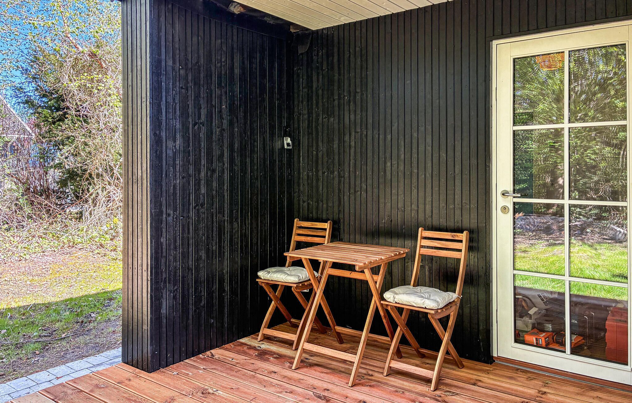 A wooden deck with two chairs and a table, a black exterior wall, and a glass door with a view of greenery.