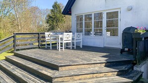 Una terraza de madera con mesa y sillas, un espacio cubierto con flores y un edificio blanco con ventanales grandes.