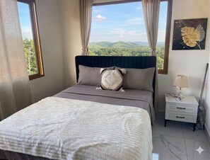 Une chambre à coucher avec un lit, une table de chevet, une lampe, un tableau et une fenêtre donnant sur des collines verdoyantes.