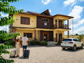 Um hotel de dois andares com uma varanda, um carro estacionado em frente e uma pessoa com malas.