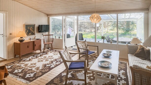 Ein gemütliches Zimmer mit Holzboden, einem Esstisch mit Stühlen, einer Couch, einem Fernseher und einem großen Fenster mit Blick ins Freie.