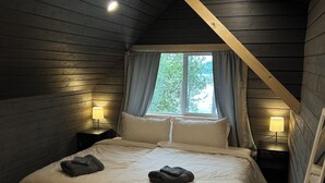 A bedroom with a wooden ceiling, a large bed with pillows and folded towels, two bedside lamps, and a window with curtains. - Lakefront Cabin on The Big Rideau (Rideau Lakes)