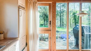 Una cocina con armarios de madera, un refrigerador y un amplio ventanal corredero que da a un espacio exterior con una parrilla y un contenedor azul.