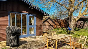 Una casa de madera con puerta azul, un patio con mesa y sillas, y una parrilla cubierta.