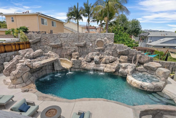 A pool with a rocky design, lounge chairs, and a hot tub.