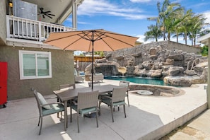 An outdoor patio with a dining table and chairs, a large umbrella, and a rock waterfall feature.
