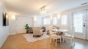 A spacious room with a dining area, a sofa, a TV mounted on the wall, and a large window with blinds.