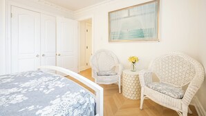 A bedroom with a bed, two wicker chairs, a small table with flowers, and a framed picture on the wall.