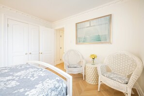 A bedroom with a bed, two wicker chairs, a small table with flowers, and a framed picture on the wall.