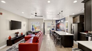 A modern kitchen with a central island, a dining area with bar stools, a red sofa, and a flat-screen TV mounted on the wall. - 4BR Near Zion -Pool, Fire Pit & Game Room (Hurricane)