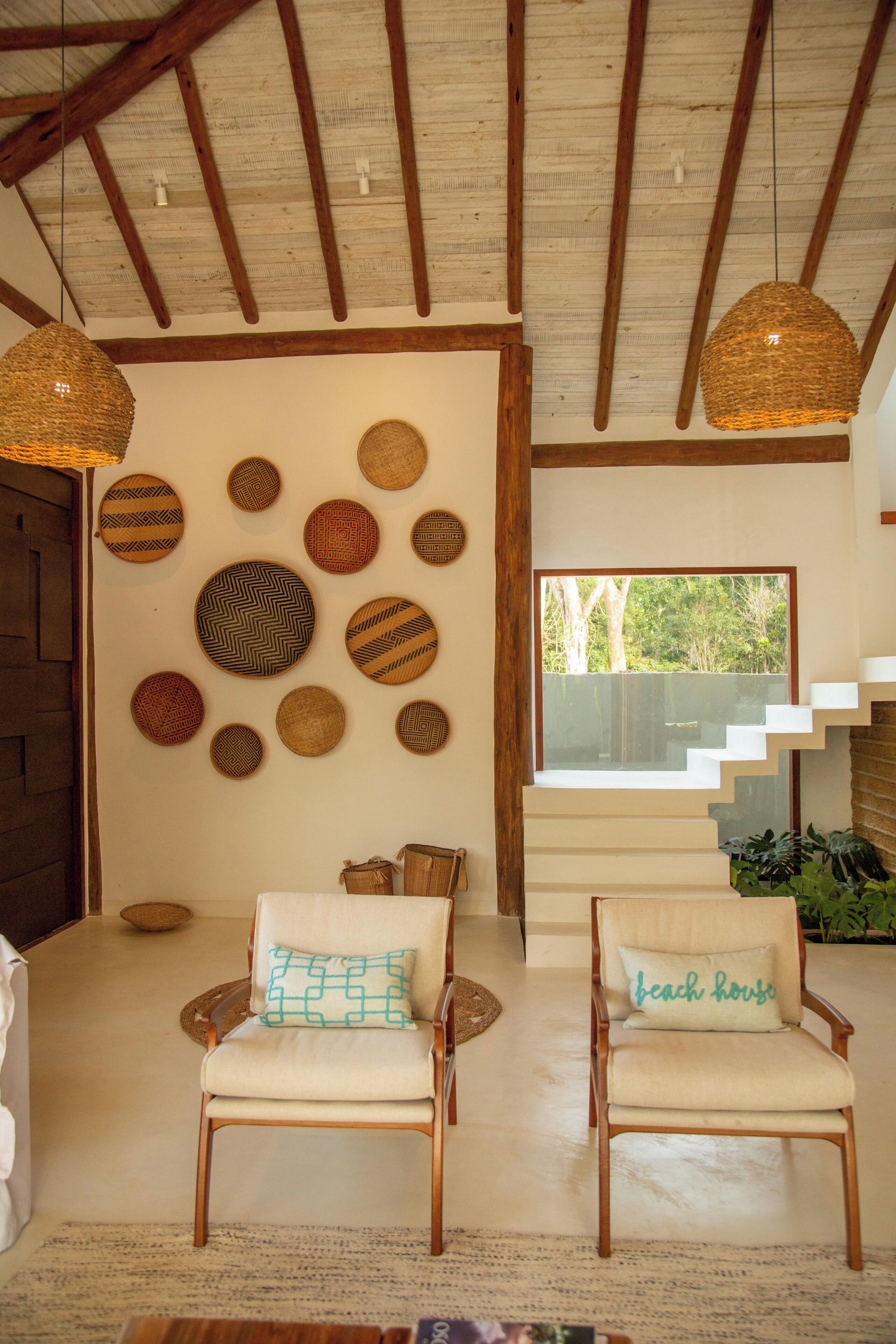 Una habitación con techo alto, vigas de madera, una escalera, dos sillones con cojines y una pared decorada con cestas tejidas.