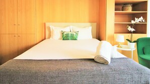 A hotel room with a bed, a green chair, a small table with a lamp, and a shelf with a plant. - Douro Apartments - Luxuryviews (Vila Nova de Gaia)