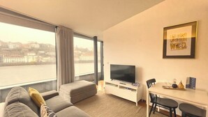 A modern hotel room with a large window, a flat-screen TV, a sofa, a dining area, and a city view. - Douro Apartments - Luxuryviews (Vila Nova de Gaia)
