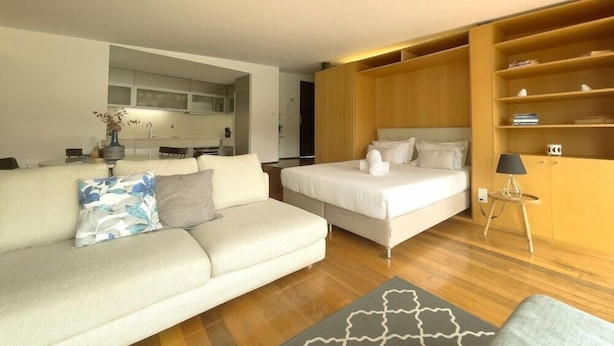A modern room with a sofa, a bed, a small table with a lamp, and a built-in wooden shelf. - Douro Apartments - Luxuryviews (Vila Nova de Gaia)
