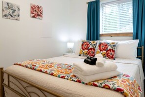 Ein ordentlich bezogenes Bett mit Blumenmuster, eine Nachttischlampe und ein Fenster mit Vorhängen.