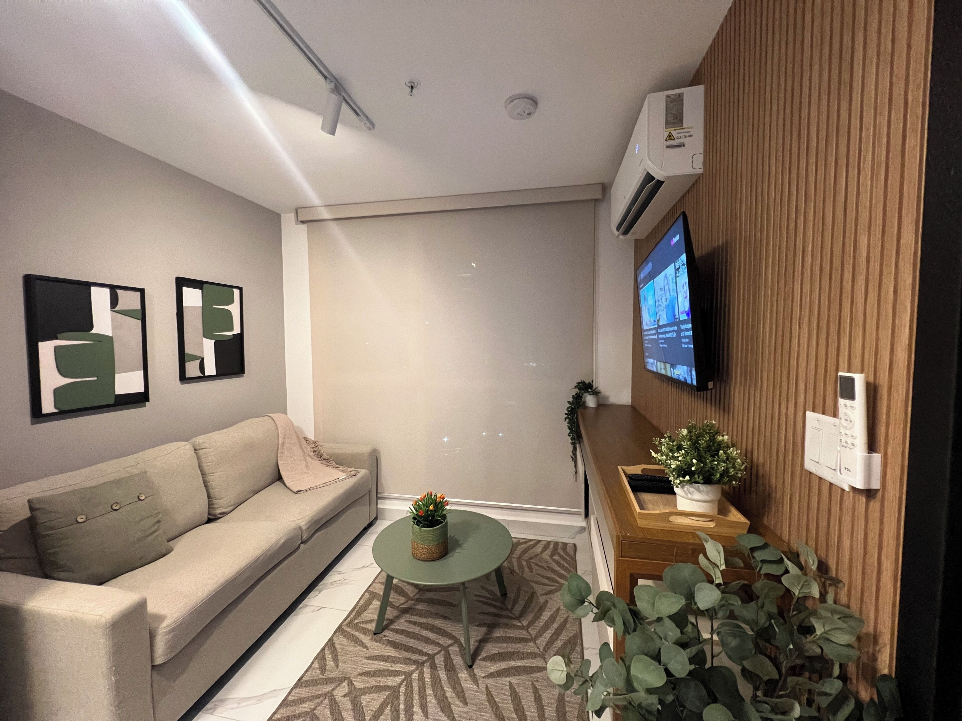 A modern living room with a sofa, a small round table with a plant, a mounted TV, and a wall-mounted air conditioner.