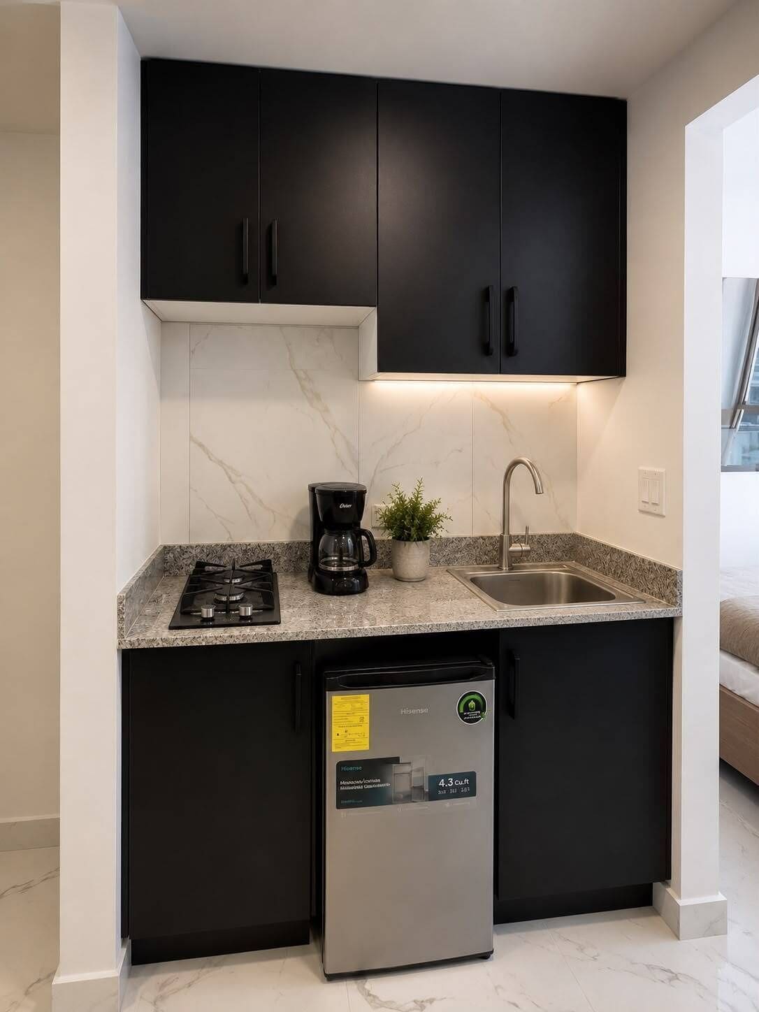 Una cocina compacta con armarios oscuros, estufa a gas, cafetera, pileta y refrigerador pequeño.